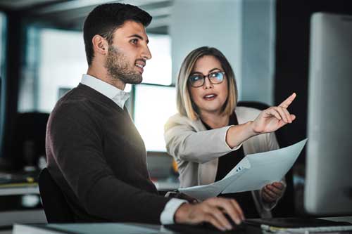 Woman who is holding papers and pointing at screen while talking to a man.