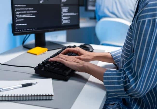 A person wearing a blue and white striped shirt types on a black keyboard in front of a computer monitor displaying code. A notebook and pen are on the desk to the left.