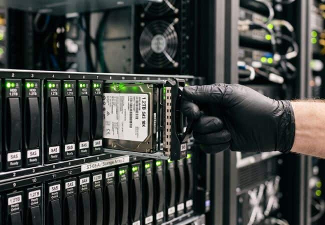a gloved hand inserts a hard drive into a server rack with multiple black drive bays and green status lights