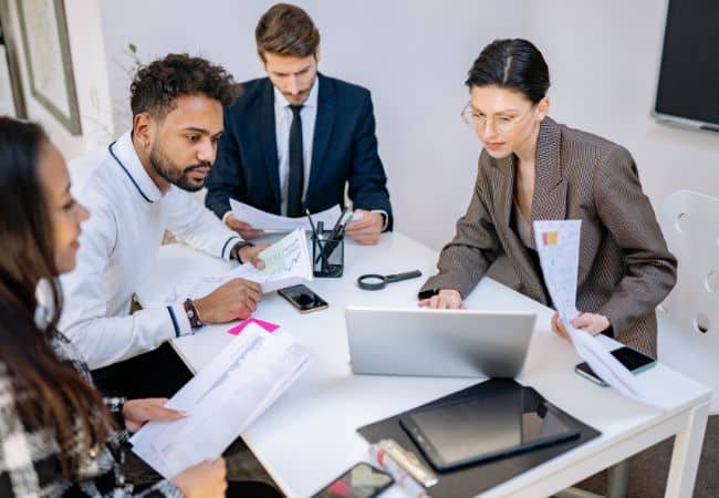 a team of professionals around a conference table reviewing documents and charts, using laptops and writing with pens while discussing ideas in an office meeting