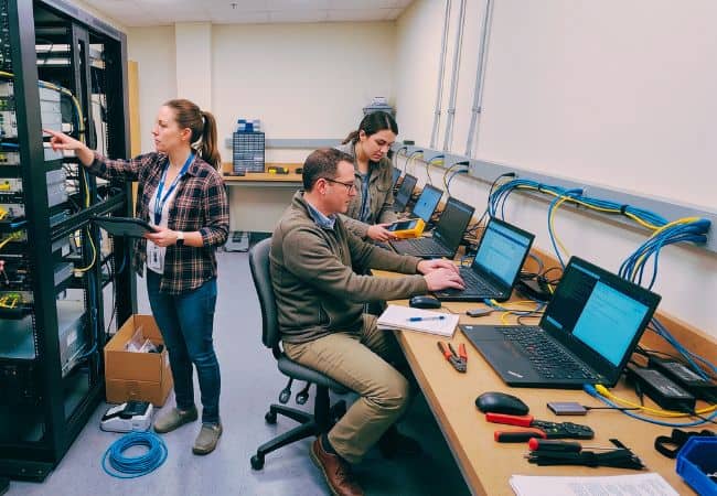 in a computer lab, three technicians work at server racks and a control console, using monitors and tools on a desk beside bundled cables and network equipment