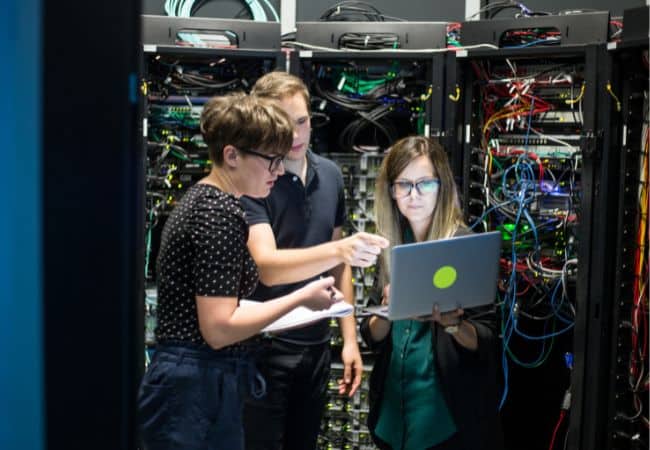 people working in front of tall server racks filled with blinking cables and network equipment, one person pointing while another checks a laptop screen