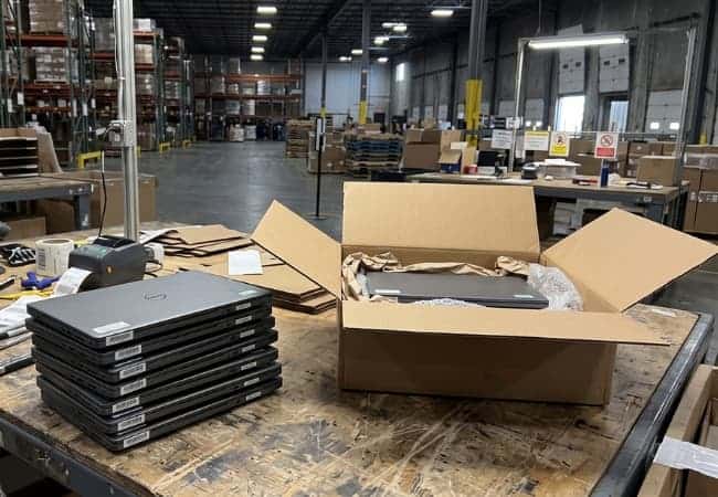 warehouse interior with stacked laptops on a worktable and open cardboard cartons filled with packing materials, shelves and loading area in the background